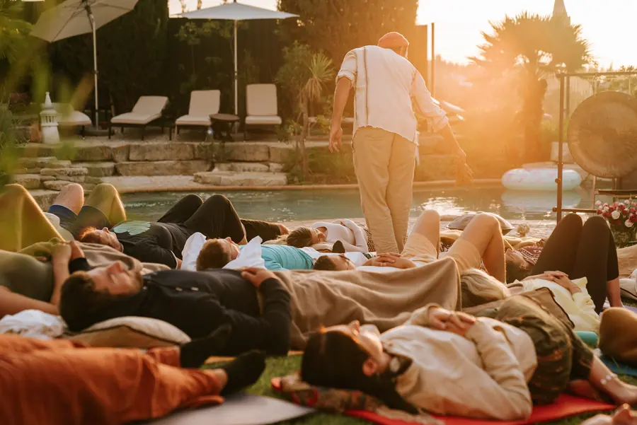 Menschen liegen bei Sonnenuntergang auf Matten neben einem Außenpool; eine Person steht in ihrer Nähe.